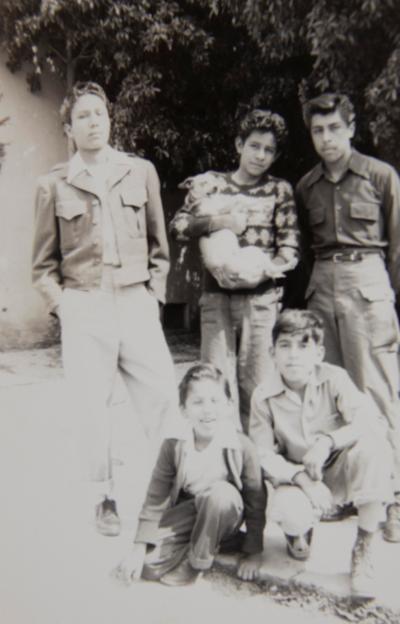 Five boys pose together. Bobby Martinez sits in the bottom left of the photograph. The other four boys are unidentified and one boy in the middle of the back row holds a dog.